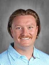 Smiling man with a mustache and medium-length hair, wearing a light blue patterned shirt, against a gray background.