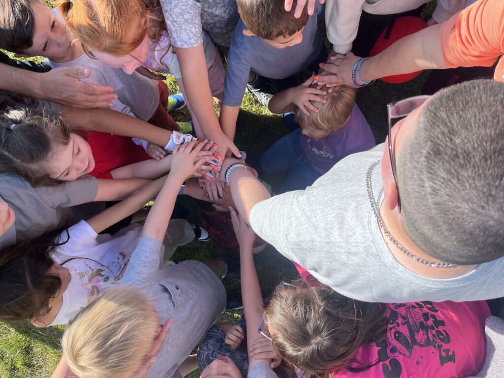 A group of children and adults gather, placing their hands together in a circle, promoting teamwork and unity.