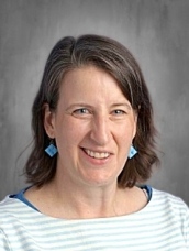 Smiling woman with shoulder-length brown hair, wearing a striped shirt and blue earrings, against a gray background.