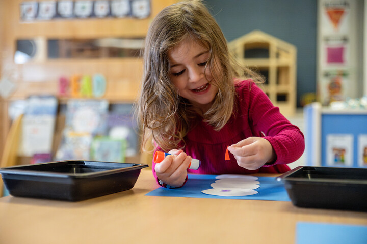 A young girl with long hair smiles while using glue and colorful paper on a craft project at a classroom table.