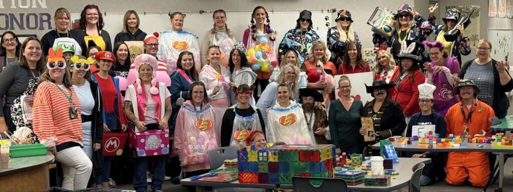 A large group of people in creative costumes, posing together in a classroom decorated with colorful toys and props.