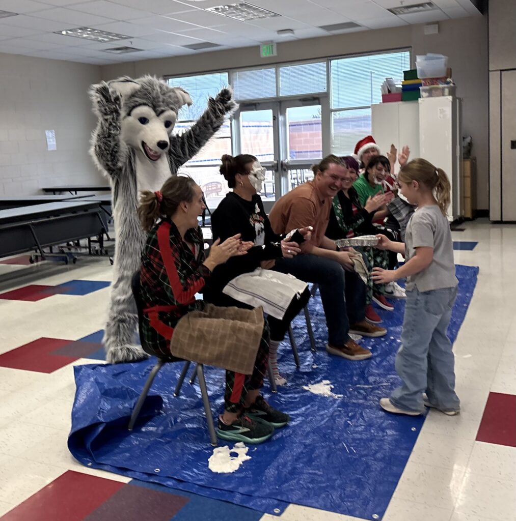 A costumed wolf mascot cheers as a child hands out gifts to seated adults in a festive cafeteria setting.