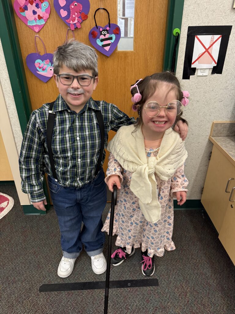 Two children dressed in playful elderly costumes stand together in front of a decorated door, smiling cheerfully.