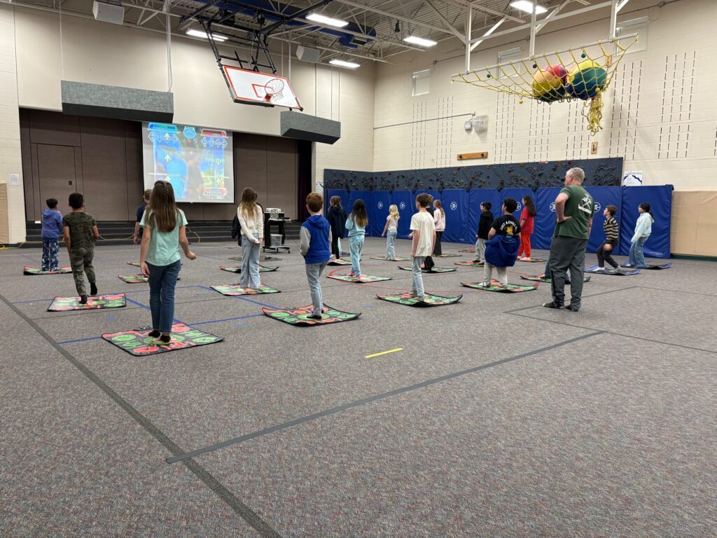 Students are lined up on mats, following a dance or movement activity guided by a projection screen in a gym.
