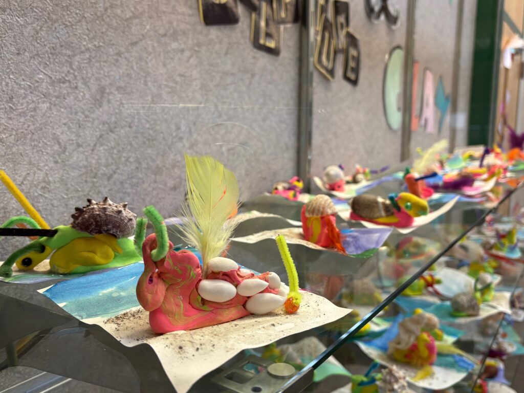 Colorful handmade snail sculptures displayed on paper, featuring various materials like clay, shells, and feathers.
