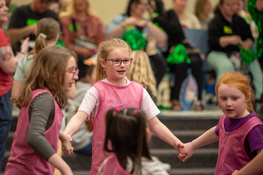 Three children in pink vests are holding hands and dancing, with a crowd of people in the background.