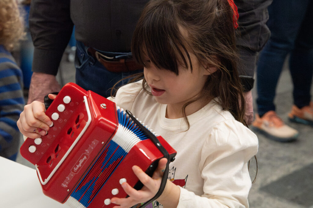 A young girl holds a small red accordion, focusing intently as she explores its buttons and keys.