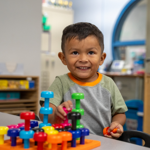 A smiling young boy plays with colorful stacking pieces on a table in a bright classroom.