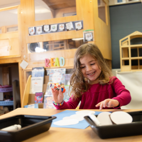 A young girl with curly hair smiles while crafting at a table, using glue and paper in a colorful classroom setting.