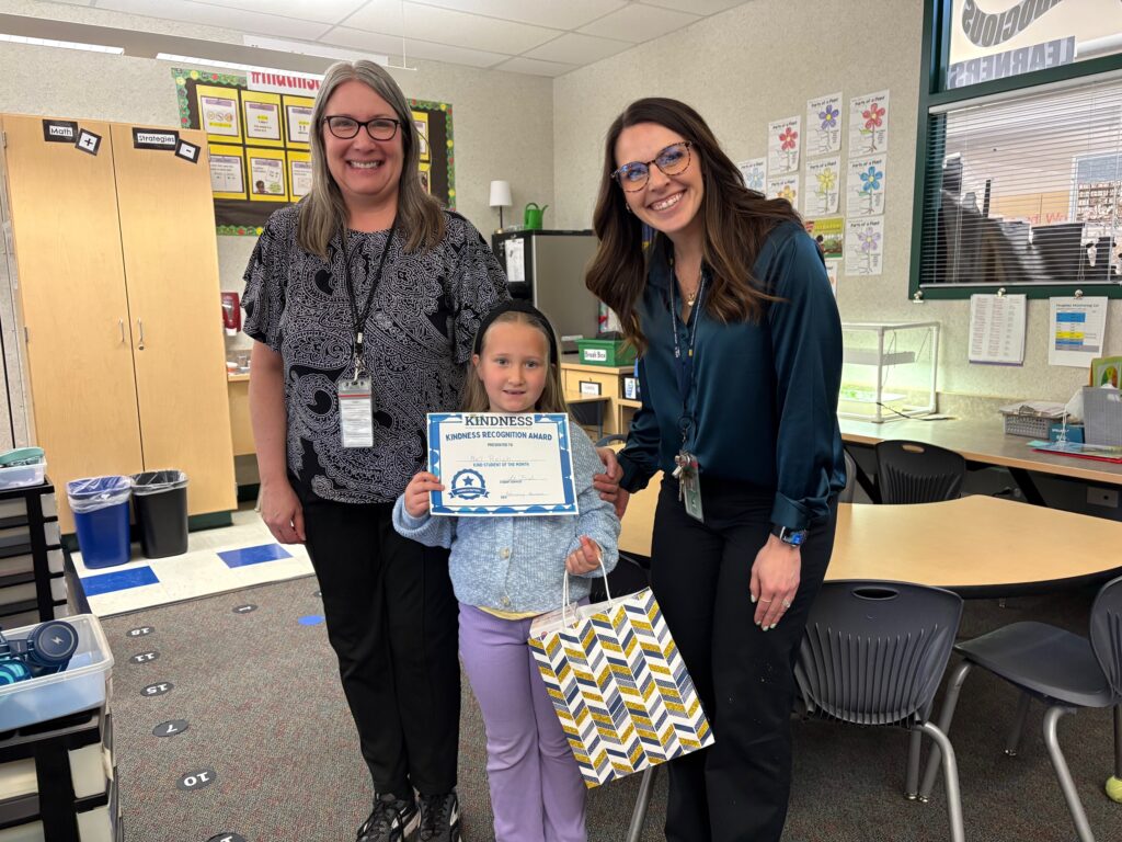 Two teachers and a young girl hold a kindness award in a classroom, surrounded by educational materials and seating.