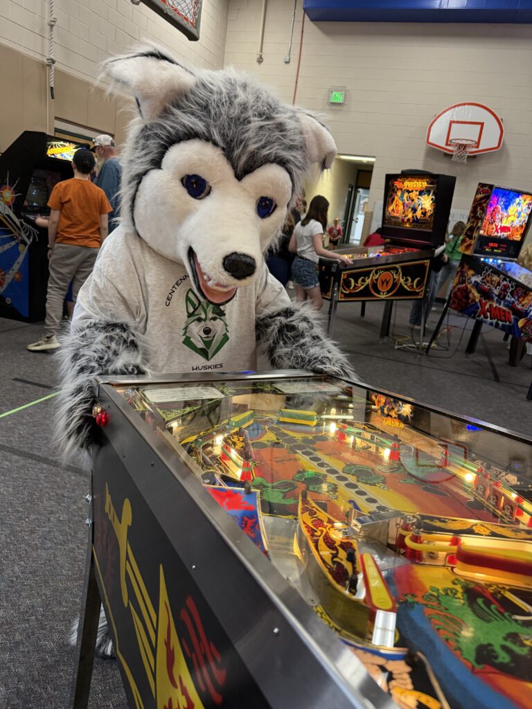 A wolf mascot in a shirt plays pinball at an event, surrounded by arcade games and people.