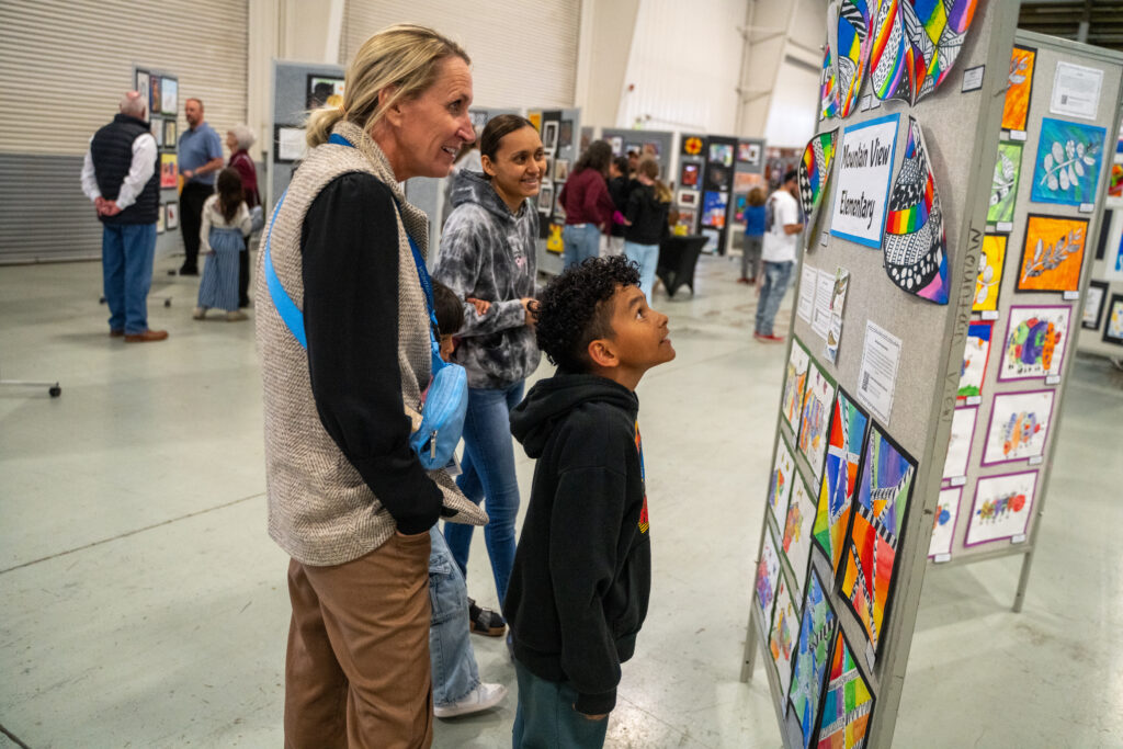 A woman and a boy admire colorful artwork at an exhibition, while others browse in the background.