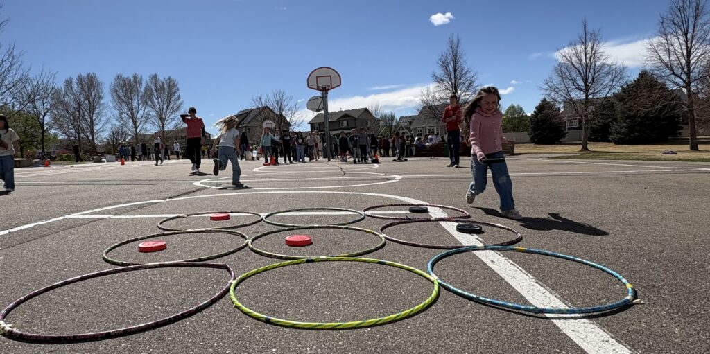 Children play outside on a sunny day, engaging in activities with hula hoops and a basketball court in the background.