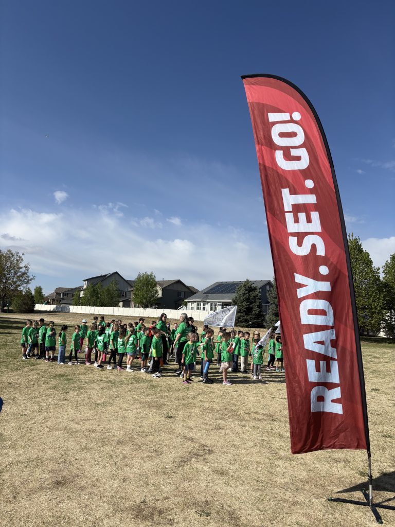 A group of children in green shirts gathers on a field under a blue sky, with a banner reading "READY. SET. GO!" nearby.
