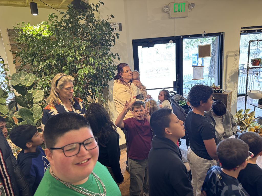 A group of children and adults gather indoors, surrounded by plants, with a door leading outside visible in the background.