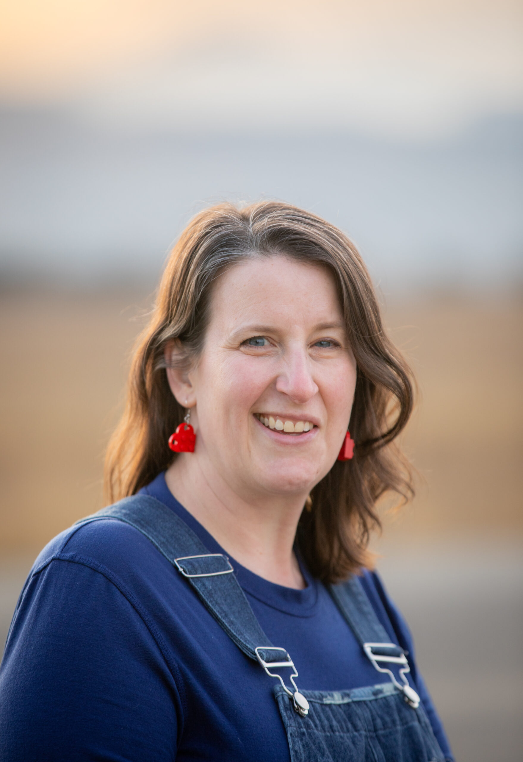 A woman with wavy hair smiles, wearing a blue shirt and denim overalls, with red strawberry earrings. Soft background.