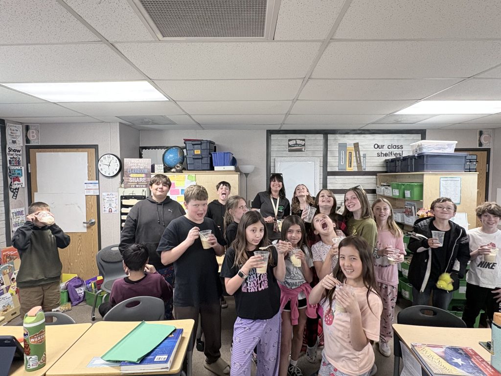 A group of students and a teacher smile and pose in a classroom, holding drinks and wearing casual clothing.