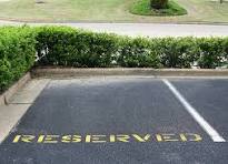 Una plaza de aparcamiento con la palabra «RESERVADO» pintada en amarillo sobre el asfalto, junto a un seto verde.